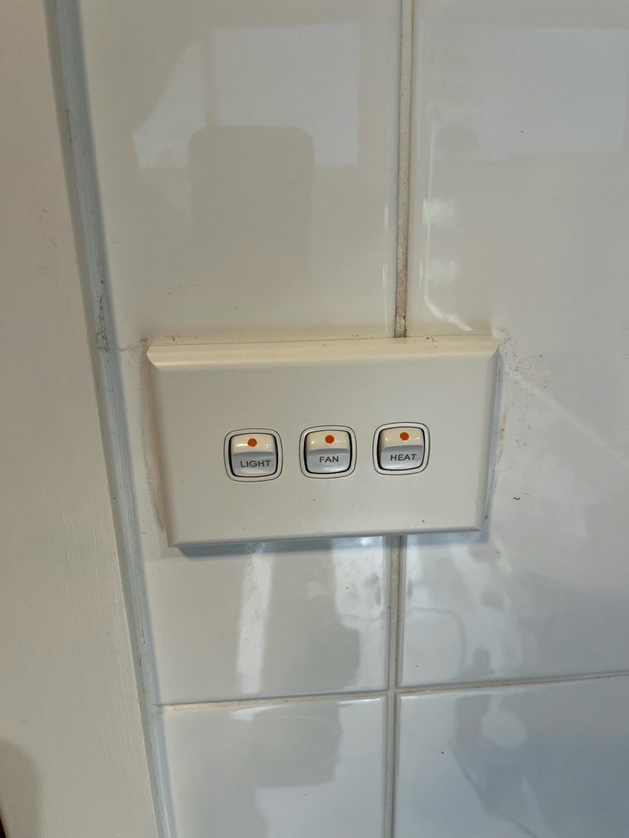 A three-gang switch plate on a tiled wall, the buttons labelled light, fan and heat