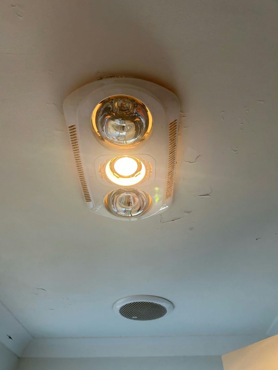 A three-in-one heat, fan and light unit with its lamps on, the ceiling paint cracked around the frame