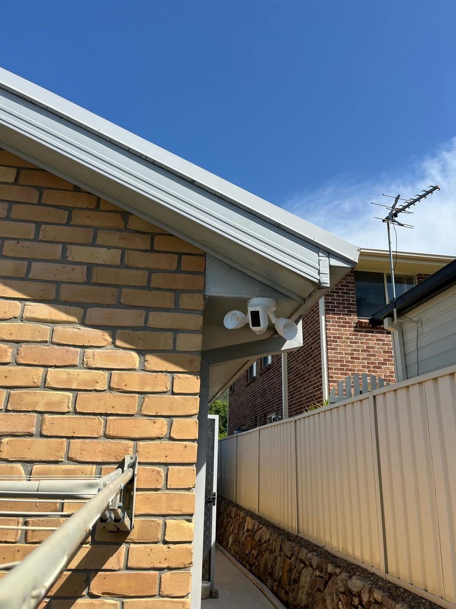 A CCTV camera mounted under the gable of a brick home, the television antenna on the ridge behind