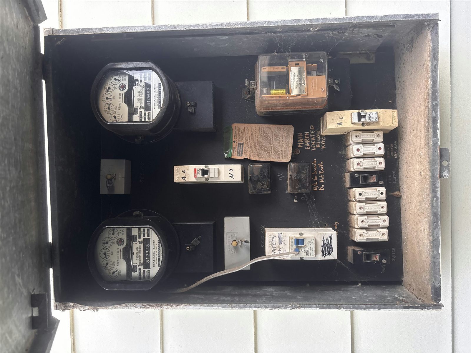 An old meter board with round analogue meters and rewireable fuses in a corroded enclosure