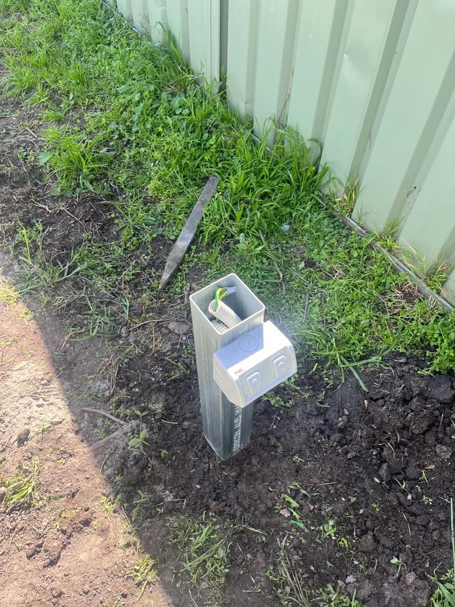 A weatherproof double outlet on a post in a freshly backfilled trench beside a fence