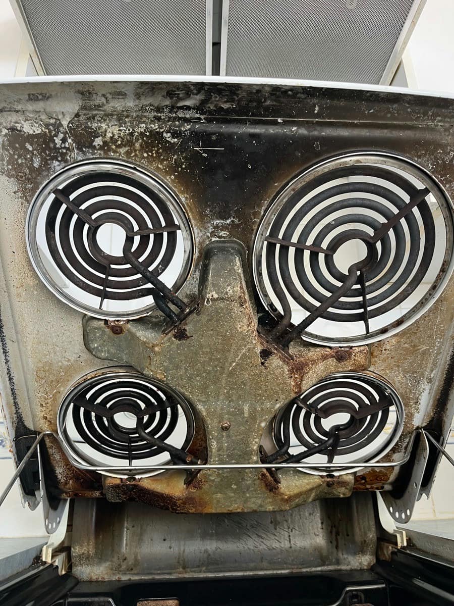 An old coil cooktop lifted to show corroded elements and a rusted pan beneath