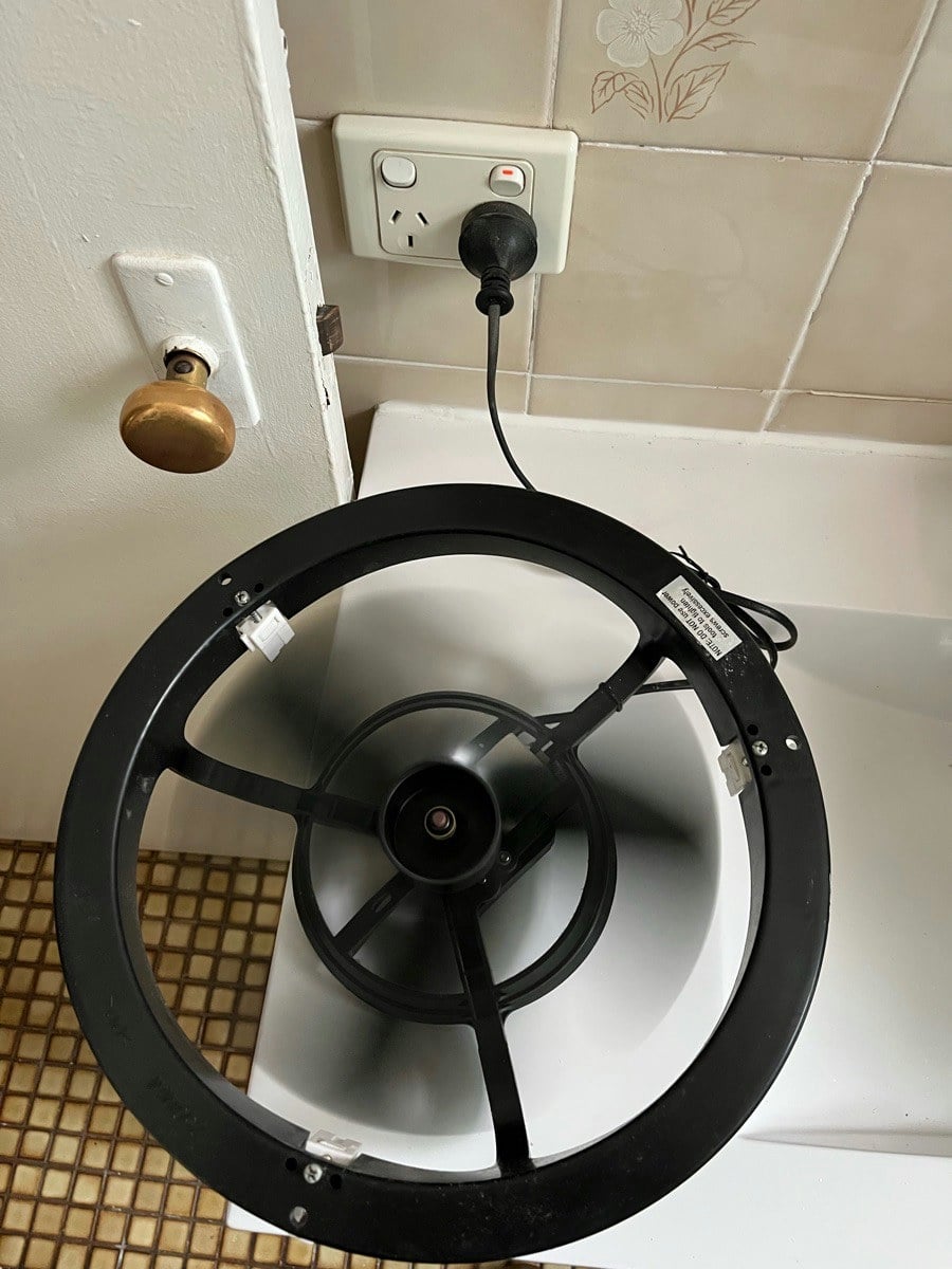 A round exhaust fan out of the ceiling with its trim ring off, resting on the bath surround